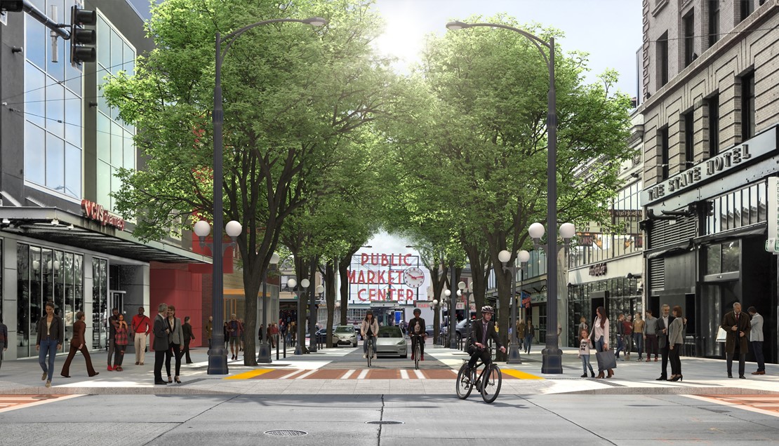 Wide sidewalks and a curbless street on the 100 Pike Street block filled with pedestrians. The street is lined with trees on both sides and people riding bicycles alongside cars with the Public Market sign in the background
